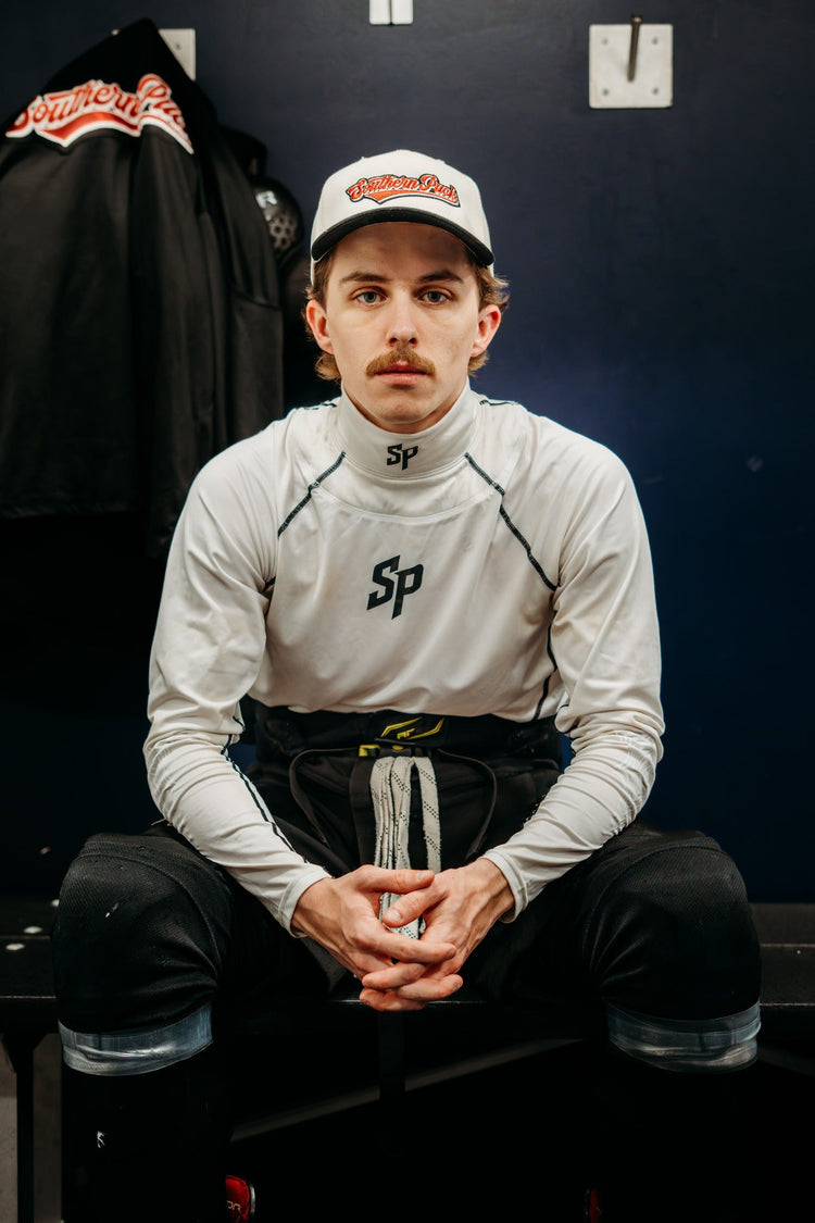 Adult athlete seated in a locker room wearing the white Southern Puck neck guard performance shirt, front view